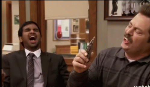 A Man In A Suit And Tie Is Laughing While Another Man Holds A Cell Phone GIF