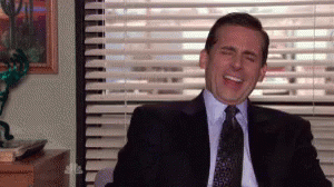 A Man In A Suit And Tie Is Laughing While Sitting In Front Of A Window With Blinds . GIF