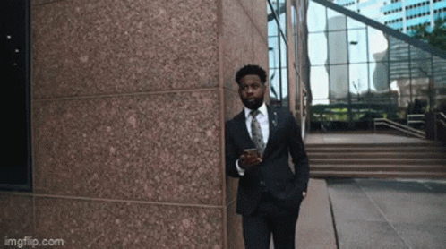A Man In A Suit And Tie Is Leaning Against A Building While Holding A Cup Of Coffee GIF
