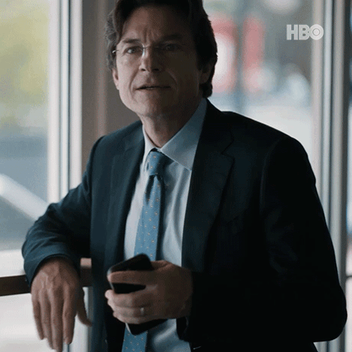A Man In A Suit And Tie Is Leaning Against A Railing Holding A Cell Phone With The Hbo Logo In The Background GIF