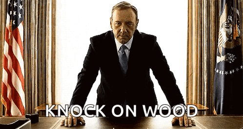 A Man In A Suit And Tie Is Leaning On A Desk With The Words " Knock On Wood " Written On It . GIF