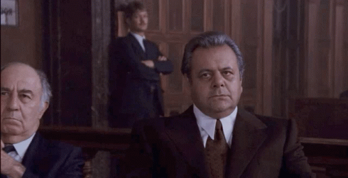 A Man In A Suit And Tie Is Looking At The Camera While Another Man Stands Behind Him . GIF