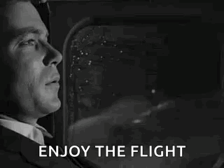 A Man In A Suit And Tie Is Looking Out The Window Of An Airplane And Saying `` Enjoy The Flight '' . GIF