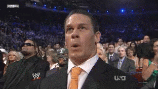 A Man In A Suit And Tie Is Making A Funny Face In Front Of A Crowd That Is Watching A Wrestling Match GIF
