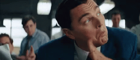 A Man In A Suit And Tie Is Making A Funny Face While Sitting In A Room With Other Men . GIF