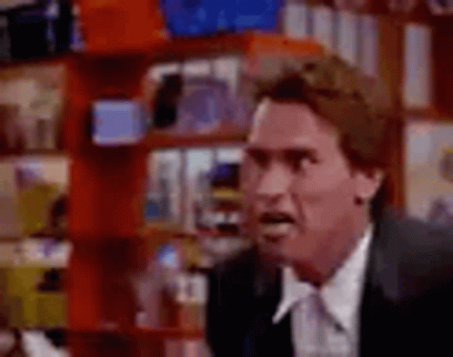 A Man In A Suit And Tie Is Making A Funny Face While Standing In Front Of A Bookshelf . GIF