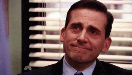 A Man In A Suit And Tie Is Making A Sad Face In Front Of A Window With Blinds . GIF
