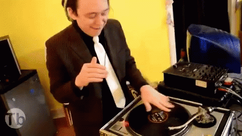 A Man In A Suit And Tie Is Playing A Record On A Turntable With The Letters Tb On The Bottom Right GIF