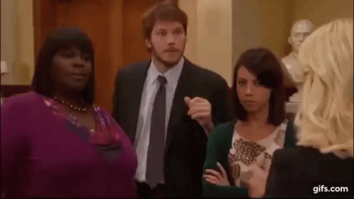 A Man In A Suit And Tie Is Pointing At A Woman In A Purple Sweater . GIF