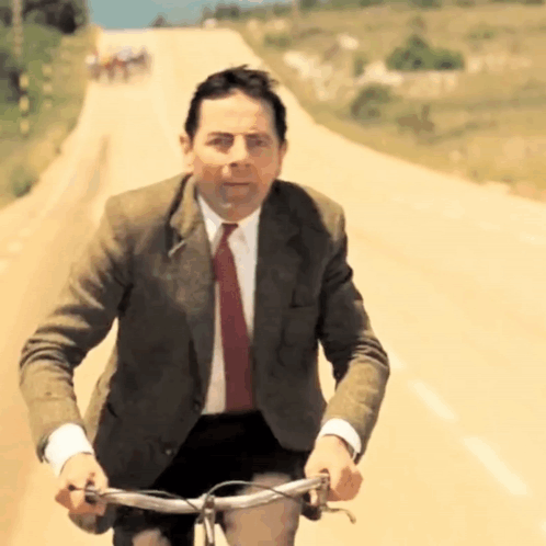 A Man In A Suit And Tie Is Riding A Bike On A Dirt Road GIF