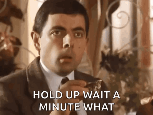 A Man In A Suit And Tie Is Saying `` Hold Up Wait A Minute What '' While Eating A Piece Of Food . GIF