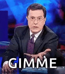 A Man In A Suit And Tie Is Sitting At A Desk And Making A Funny Face While Saying Gimme . GIF