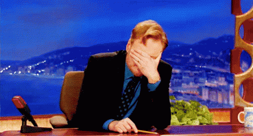 A Man In A Suit And Tie Is Sitting At A Desk Covering His Face With His Hand . GIF