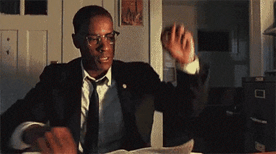 A Man In A Suit And Tie Is Sitting At A Desk Holding A Piece Of Paper . GIF