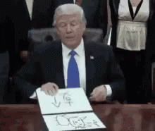 A Man In A Suit And Tie Is Sitting At A Desk Holding A Piece Of Paper GIF
