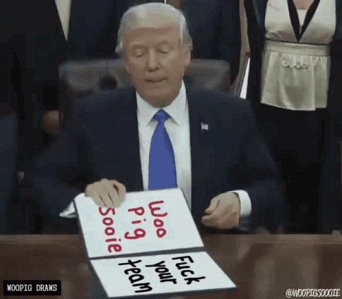 A Man In A Suit And Tie Is Sitting At A Desk Holding A Sign That Says " Whoo Pig Sooie Fuck Your Team " GIF
