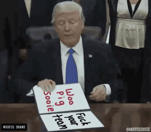 A Man In A Suit And Tie Is Sitting At A Desk Holding A Sign That Says " Woo Pig Sooie Fuck Your Team " GIF