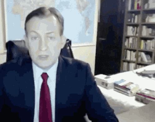 A Man In A Suit And Tie Is Sitting At A Desk In A Library . GIF
