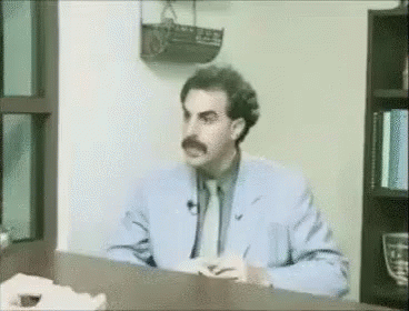 A Man In A Suit And Tie Is Sitting At A Desk In Front Of A Bookshelf . GIF