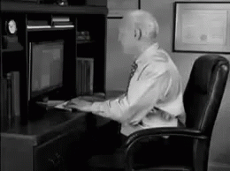 A Man In A Suit And Tie Is Sitting At A Desk In Front Of A Computer Monitor . GIF