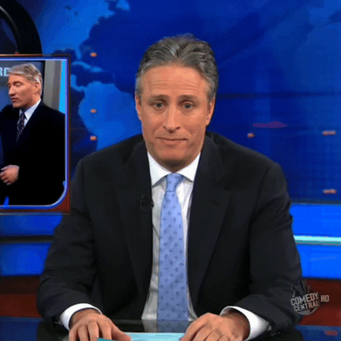 A Man In A Suit And Tie Is Sitting At A Desk In Front Of A Screen That Says Comedy Central Hd GIF
