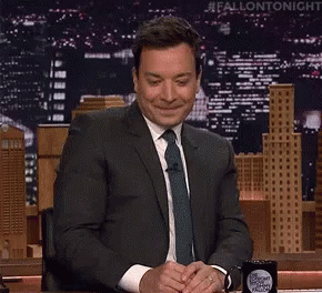A Man In A Suit And Tie Is Sitting At A Desk In Front Of A Sign That Says Fallon Tonight GIF