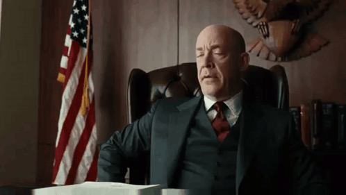 A Man In A Suit And Tie Is Sitting At A Desk In Front Of An American Flag . GIF