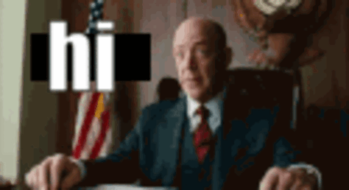 A Man In A Suit And Tie Is Sitting At A Desk In Front Of An American Flag With The Word Hi Above Him GIF