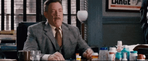 A Man In A Suit And Tie Is Sitting At A Desk With A Bunch Of Pills On It . GIF