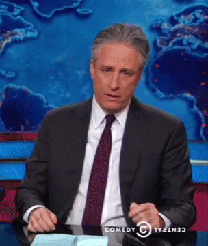 A Man In A Suit And Tie Is Sitting At A Desk With A Comedy Central Logo Behind Him GIF