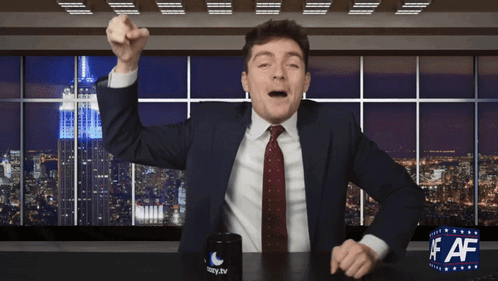 A Man In A Suit And Tie Is Sitting At A Desk With A Cup That Says Lazy.tv On It GIF