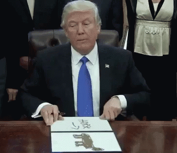 A Man In A Suit And Tie Is Sitting At A Desk With A Piece Of Paper That Says ' Trump ' On It GIF