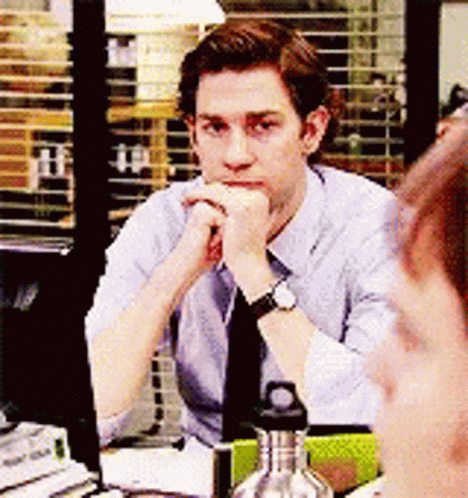 A Man In A Suit And Tie Is Sitting At A Desk With His Hand On His Chin . GIF