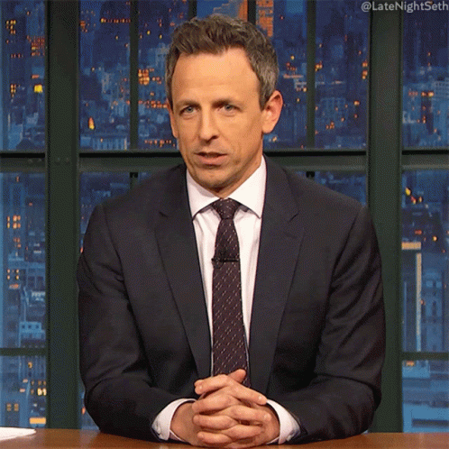 A Man In A Suit And Tie Is Sitting At A Desk With His Hands Folded GIF