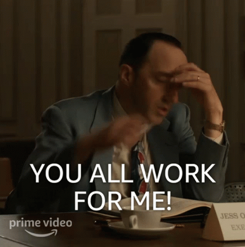 A Man In A Suit And Tie Is Sitting At A Desk With His Hands In The Air And Says You All Work For Me GIF