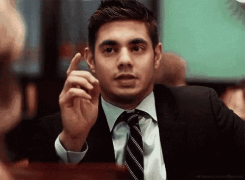 a man in a suit and tie is sitting at a table and pointing his finger .