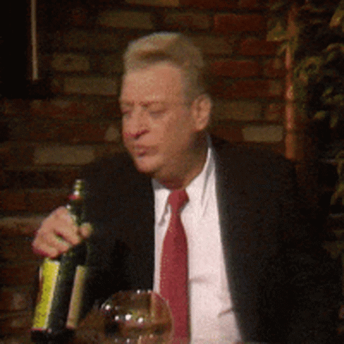 A Man In A Suit And Tie Is Sitting At A Table Holding A Bottle Of Wine GIF