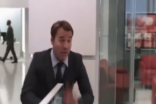 A Man In A Suit And Tie Is Sitting At A Table In A Room With A Glass Wall . GIF