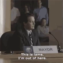 A Man In A Suit And Tie Is Sitting At A Table In Front Of A Microphone And A Sign That Says Mayor . GIF
