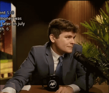 A Man In A Suit And Tie Is Sitting At A Table In Front Of A Microphone With A Mug That Says Af On It GIF