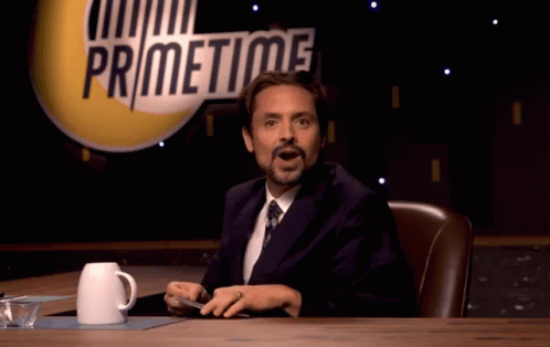 A Man In A Suit And Tie Is Sitting At A Table In Front Of A Sign That Says Primetime GIF