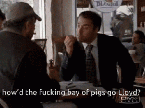 A Man In A Suit And Tie Is Sitting At A Table Talking To Another Man In A Hat . GIF