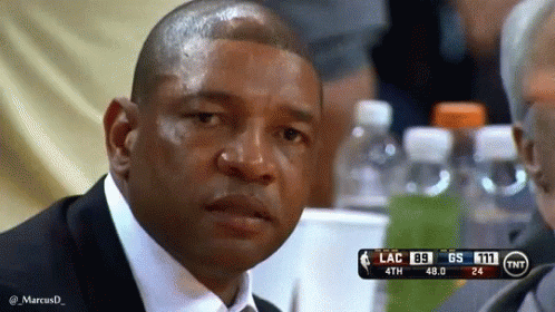 A Man In A Suit And Tie Is Sitting At A Table Watching A Basketball Game With A Scoreboard Behind Him . GIF