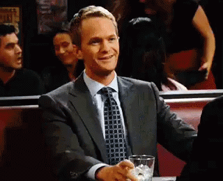A Man In A Suit And Tie Is Sitting At A Table With A Drink . GIF