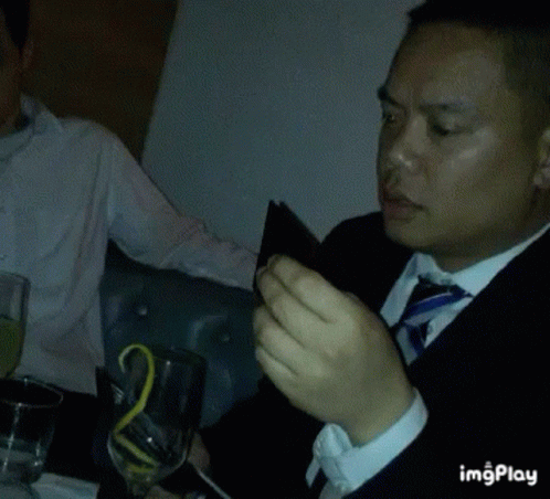 A Man In A Suit And Tie Is Sitting At A Table With A Glass Of Wine And A Wallet . GIF