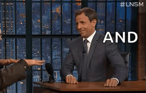 A Man In A Suit And Tie Is Sitting At A Table With A Microphone And Talking To Another Man . GIF