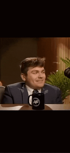 A Man In A Suit And Tie Is Sitting At A Table With A Mug That Says ' Af ' On It GIF