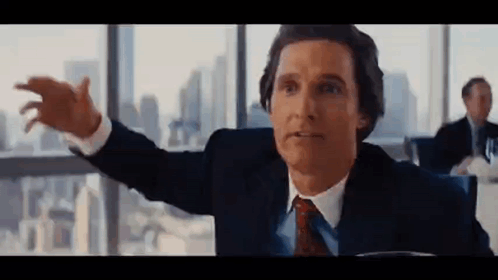 A Man In A Suit And Tie Is Sitting At A Table With His Arms Outstretched In Front Of A Window . GIF