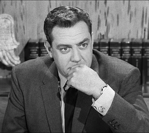 A Man In A Suit And Tie Is Sitting At A Table With His Hand On His Chin And Looking At The Camera . GIF