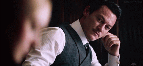 A Man In A Suit And Tie Is Sitting At A Table With His Hands On His Chin . GIF